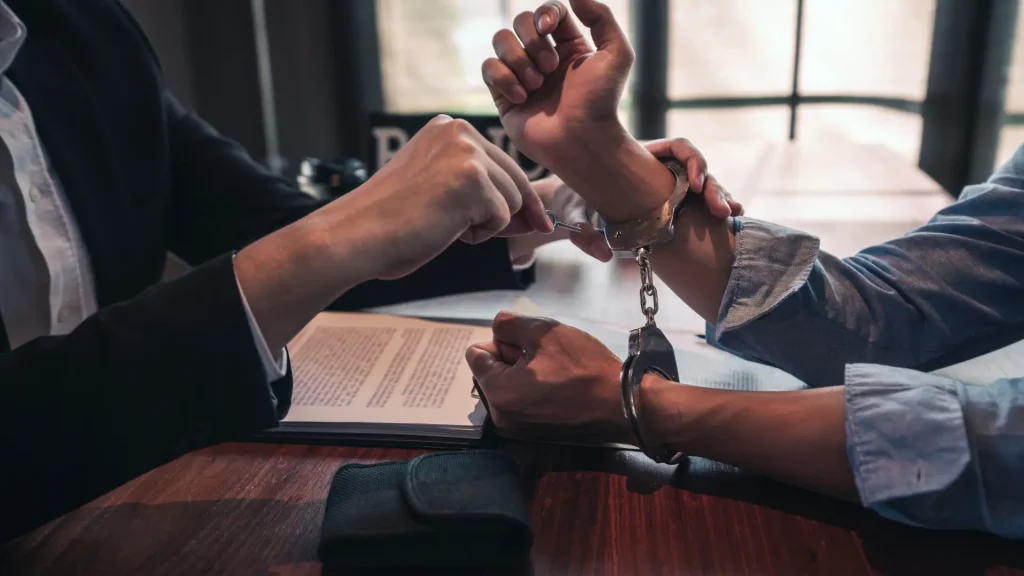 Two hands being handcuffed by an officer with a document on the table in the background.